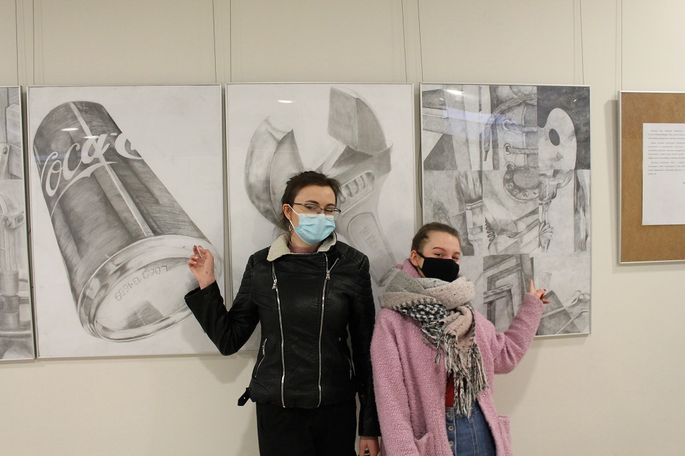 Studentki w maseczkach na twarzy przy ścianie, na której wiszą prace rysunku